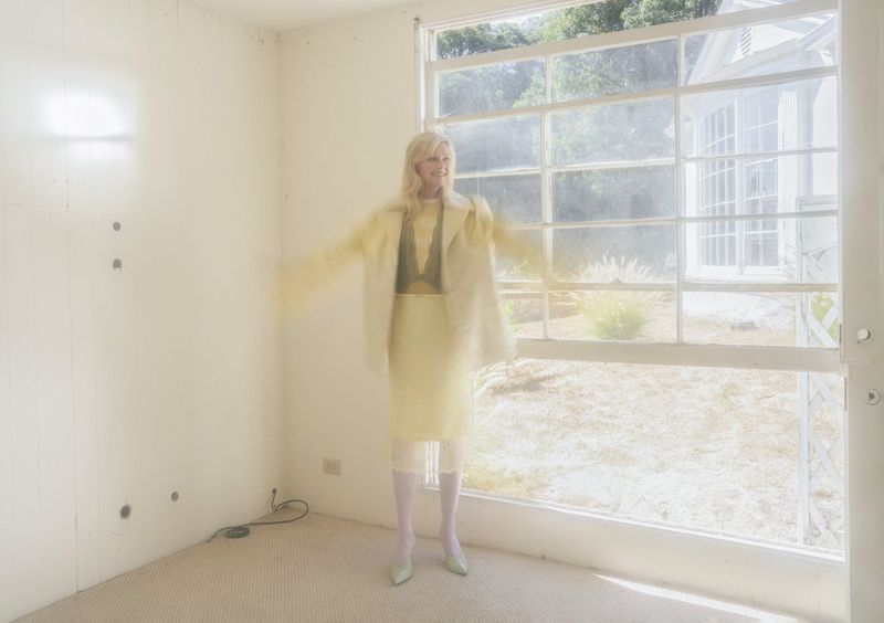 Adult female in a yellow outfit standing in an empty room with a large window and blurred motion, bathed in bright, diffused light.