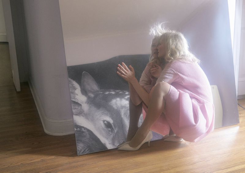 Woman in pink dress crouches next to a large monochrome painting of a fawn in an indoor setting with wooden floors.