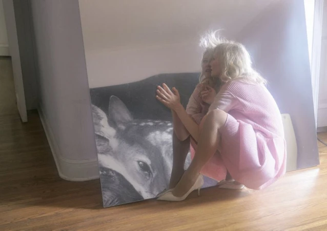 Woman in pink dress crouches next to a large monochrome painting of a fawn in an indoor setting with wooden floors.