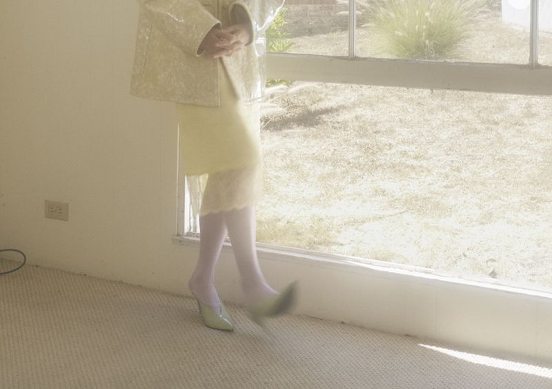 Woman in light yellow dress and cream jacket, wearing green heels, stands in a room with a window overlooking a grassy area.