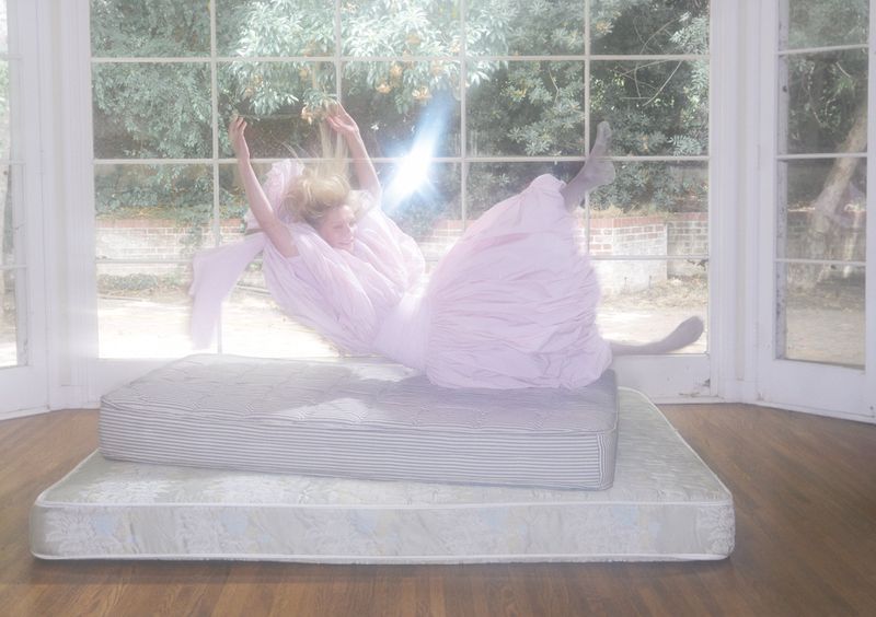 Woman in a pink dress floats above mattresses in an indoor setting, with a window and greenery in the background, creating a surreal image.
