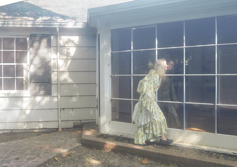 Woman in a floral dress gazes out a large window of a white house, reflecting her image, in a brightly lit outdoor scene.