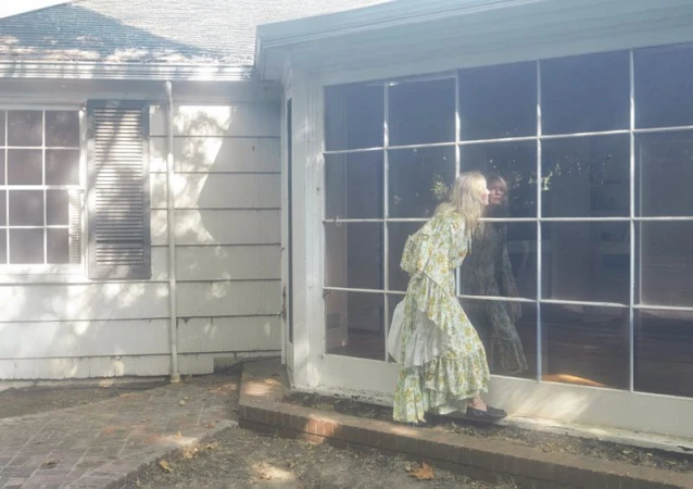 Woman in a floral dress gazes out a large window of a white house, reflecting her image, in a brightly lit outdoor scene.