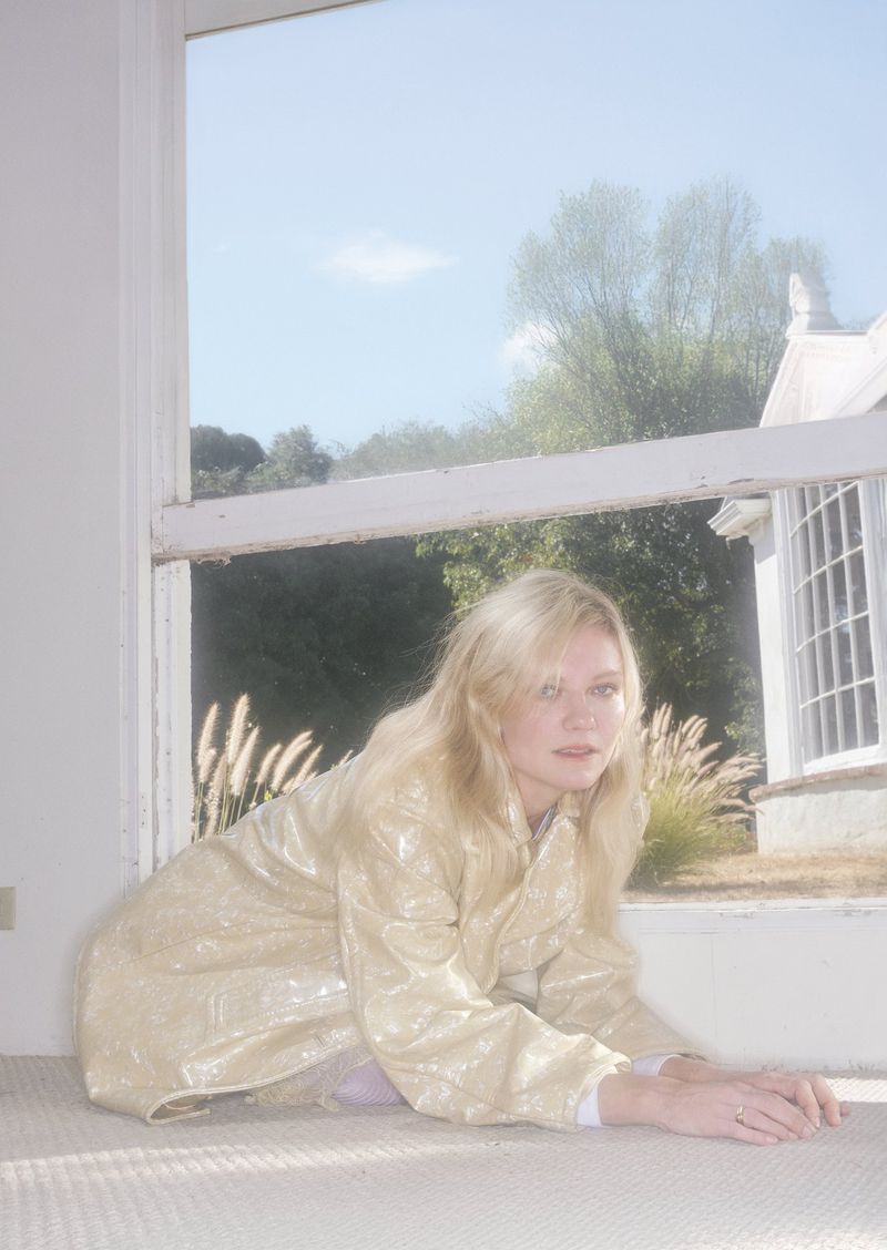 Blonde woman in a shimmering beige coat posing on floor in front of a window with an outdoor view of grass and white building.