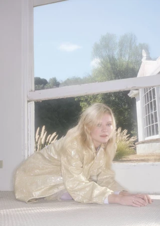 Blonde woman in a shimmering beige coat posing on floor in front of a window with an outdoor view of grass and white building.