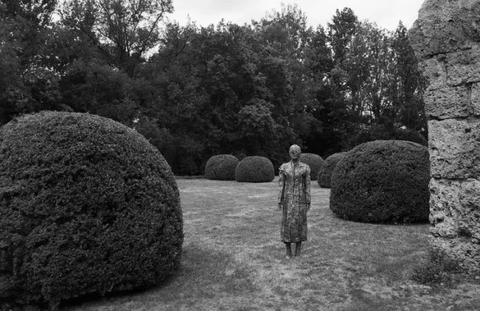 Monochrome shot of a female sculpture standing in a grassy garden surrounded by manicured shrubs and a partial stone wall.