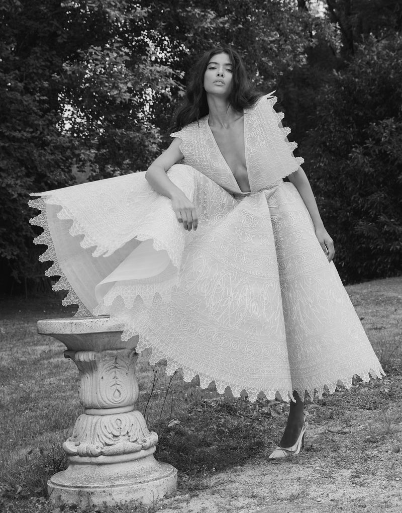 Woman in an ornate white dress poses in a garden next to an ornate stone pedestal with trees in the background in a black and white image.