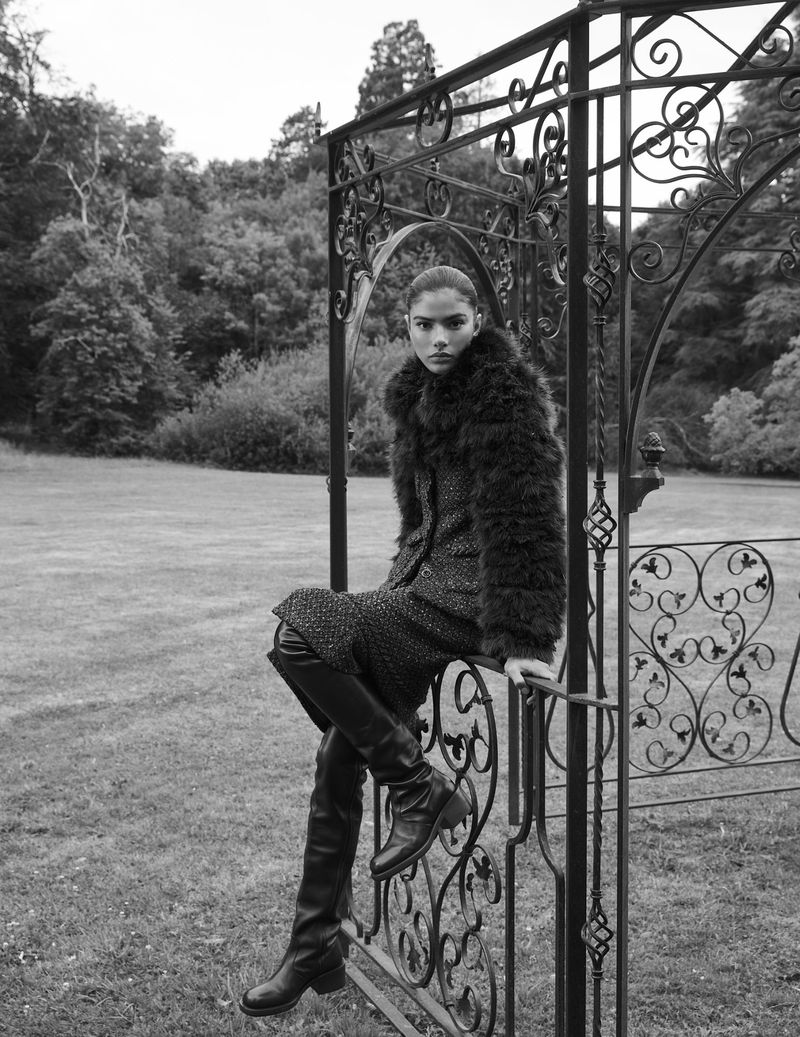 Monochrome portrait of a woman in a fur-collared coat and high boots sitting on a decorative metal fence in a park-like setting.