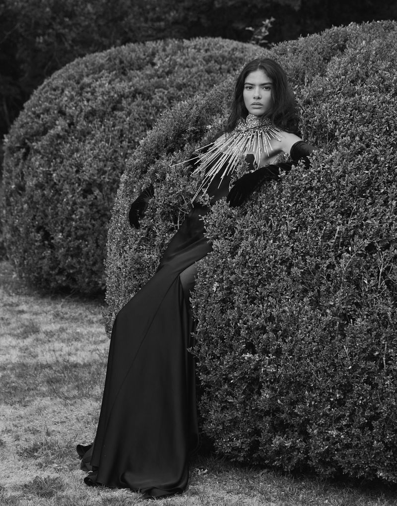 A woman in a long black dress and gloves is posing in a garden, surrounded by round bushes. The photo is black and white.