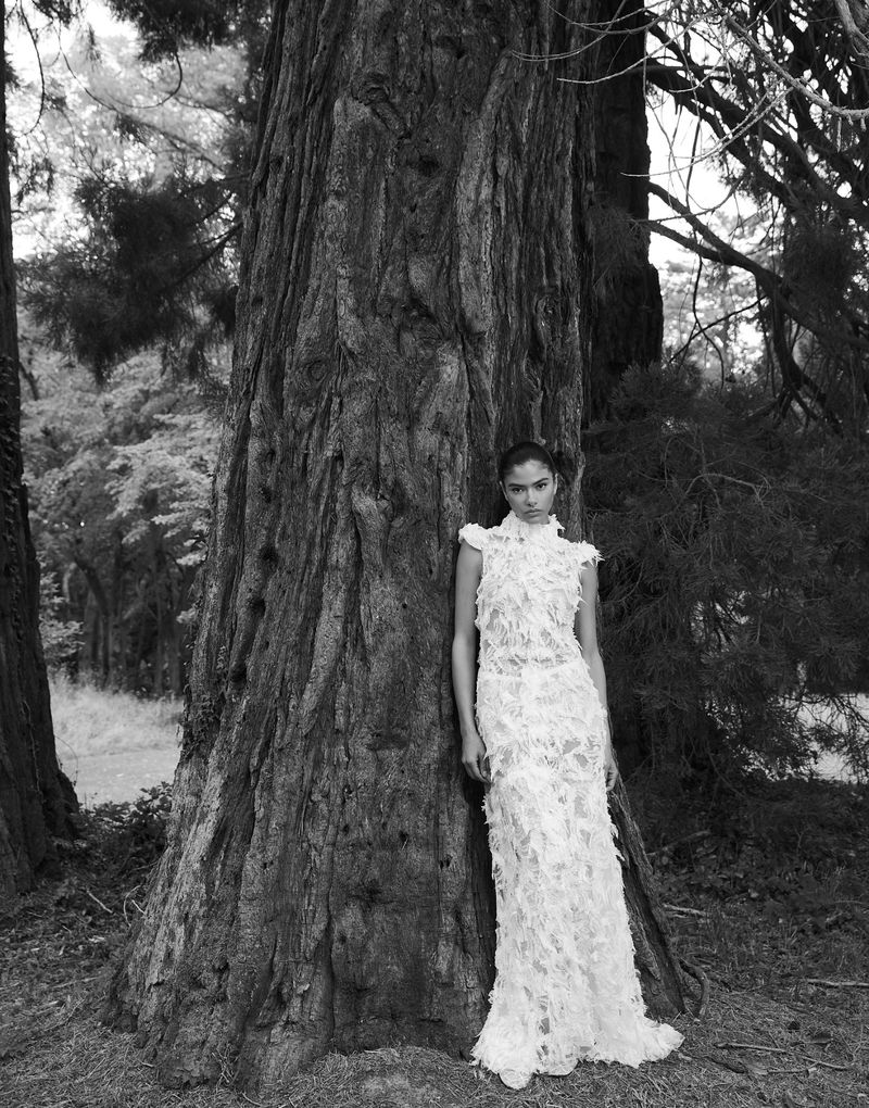 Woman in an elaborate gown poses next to a large tree in a forest setting, creating a striking black and white fashion shot.