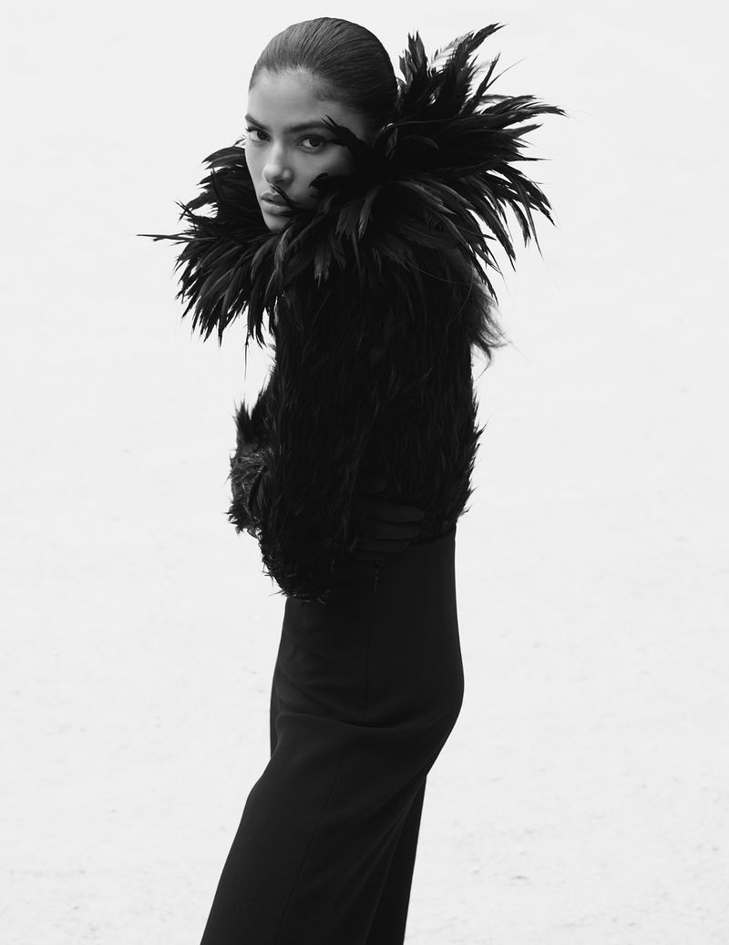 Woman in a black dress and feathered jacket poses against a white background in a studio shot.