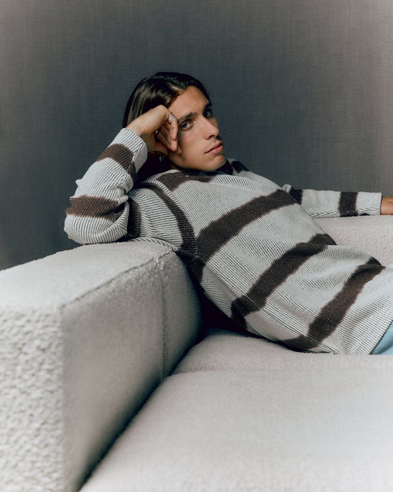 Young man with long hair relaxes on a light-colored couch, wearing a striped sweater in a casual indoor setting with soft lighting.