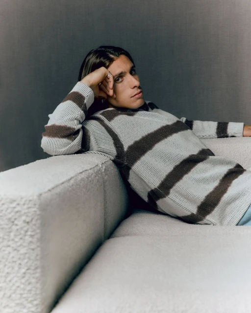 Young man with long hair relaxes on a light-colored couch, wearing a striped sweater in a casual indoor setting with soft lighting.