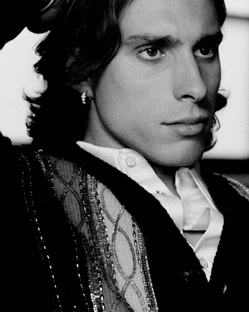 Portrait of a dark-haired adult male with an earring, wearing a decorative cardigan over a collared shirt, in a close-up black and white shot.