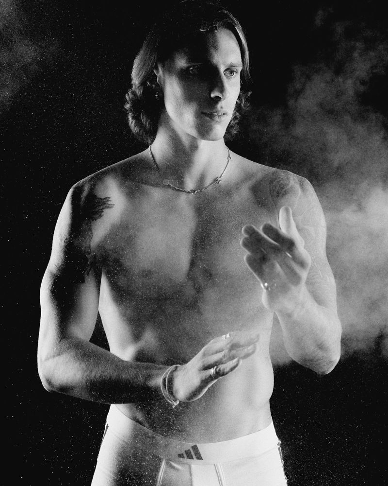 Shirtless adult male with long brown hair wearing athletic shorts, jewelry, and dust particles against a dark background.