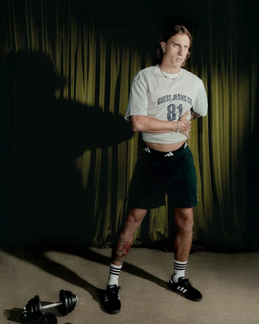 Full-body shot of an adult male in a Guess t-shirt and Adidas shorts, standing in front of gold curtains, with weights on the floor.