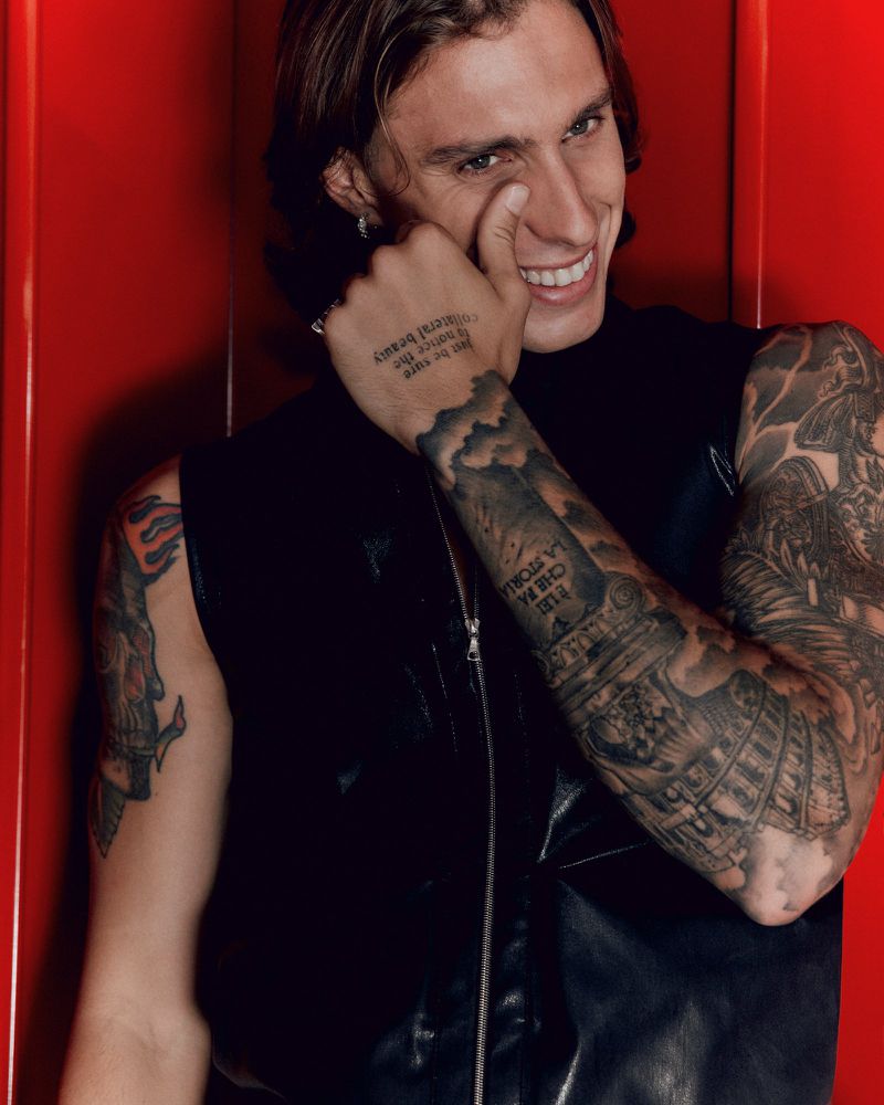 Smiling tattooed adult male portrait in a black vest against a red wall, showing handwritten text on his hand and detailed arm tattoos.