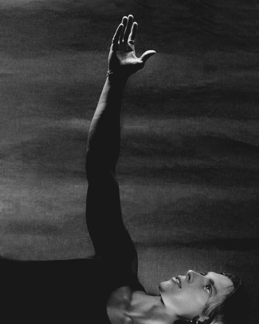 Black and white portrait of a shirtless adult male lying down with his arm extended upwards against a textured background.