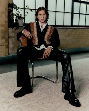 Riccardo Calafiori seated on a chair wearing a patterned knit cardigan and black leather trousers for Versus editorial.