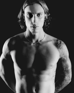 Portrait of Riccardo Calafiori shirtless with long hair and tattoos wearing a silver necklace against a black background.