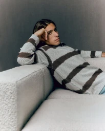 Riccardo Calafiori wearing a striped sweater, reclining on a textured white sofa against a grey wall.