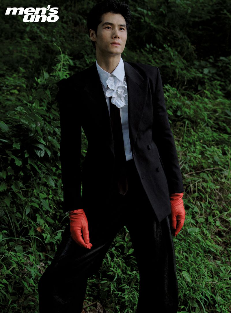 Asian male model in black suit, white shirt, and red gloves poses in a lush green outdoor setting for fashion editorial.