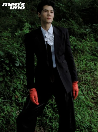 Asian male model in black suit, white shirt, and red gloves poses in a lush green outdoor setting for fashion editorial. 