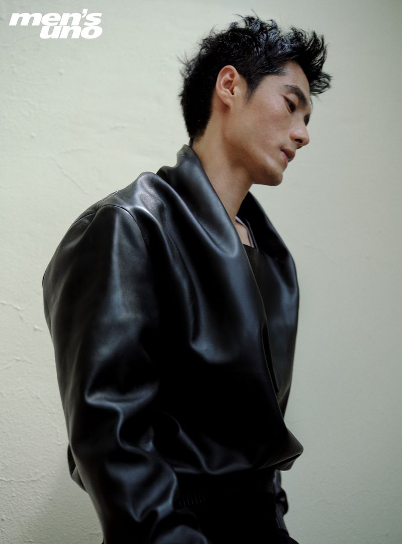 Profile portrait of a young adult male with dark, wet hair, wearing a shiny black leather jacket, against a light neutral wall indoors.