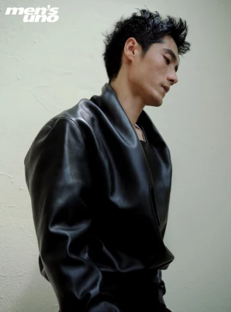 Profile portrait of a young adult male with dark, wet hair, wearing a shiny black leather jacket, against a light neutral wall indoors.