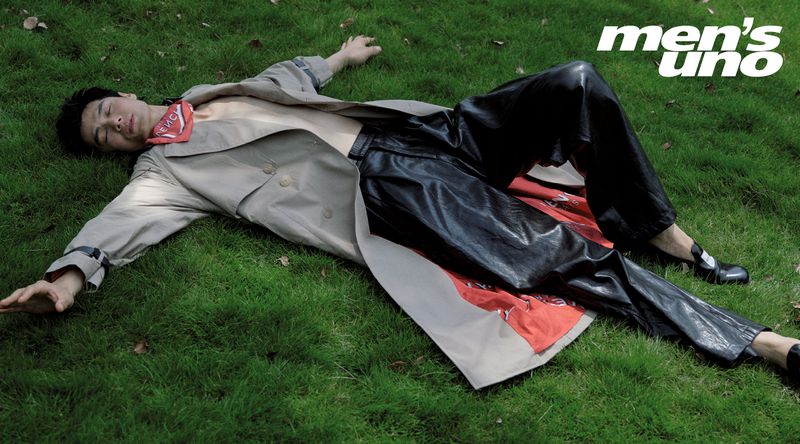 Adult man lies on green grass wearing a tan trench coat and black leather pants in a fashion editorial with magazine logo.