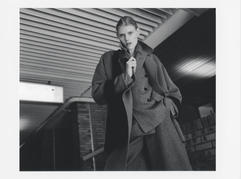 Monochromatic fashion shot of a young woman in a wool coat and suit. Indoor setting with a unique ceiling design. Editorial style photography.