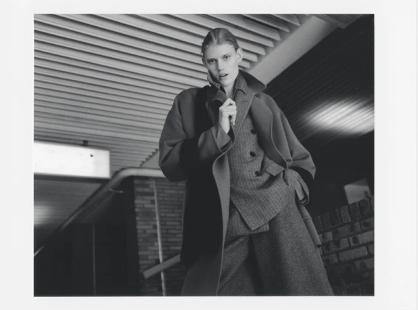 Monochromatic fashion shot of a young woman in a wool coat and suit. Indoor setting with a unique ceiling design. Editorial style photography.