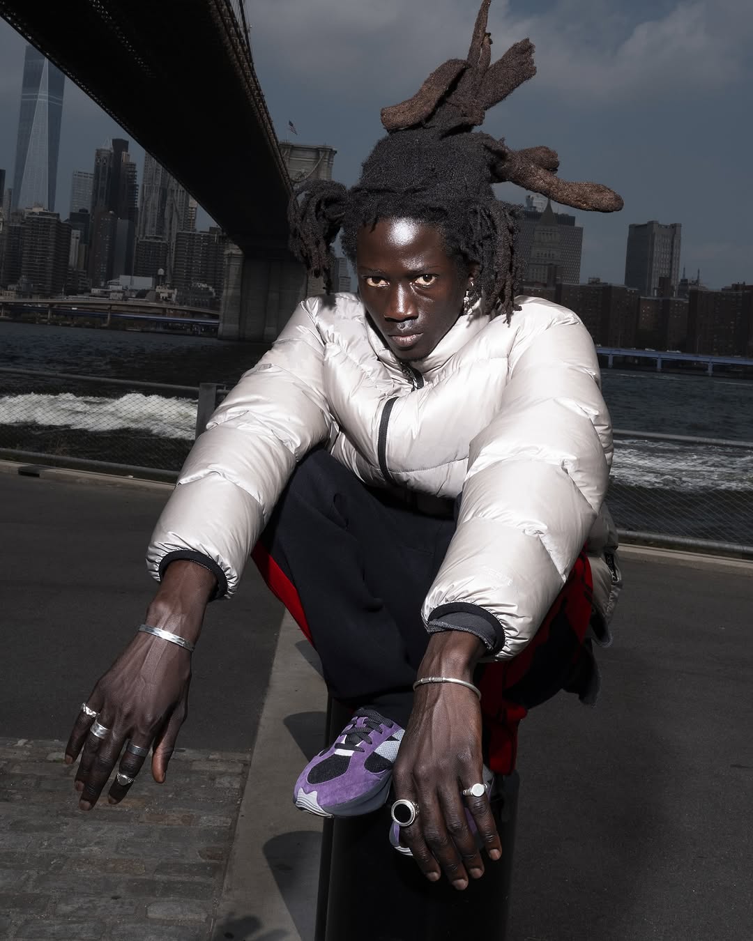 Adult male with dreadlocks sits against a backdrop of the Brooklyn Bridge and NYC skyline, wearing a silver puffer jacket and sneakers.