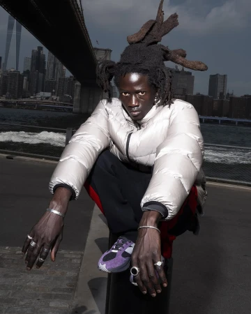 Adult male with dreadlocks sits against a backdrop of the Brooklyn Bridge and NYC skyline, wearing a silver puffer jacket and sneakers.