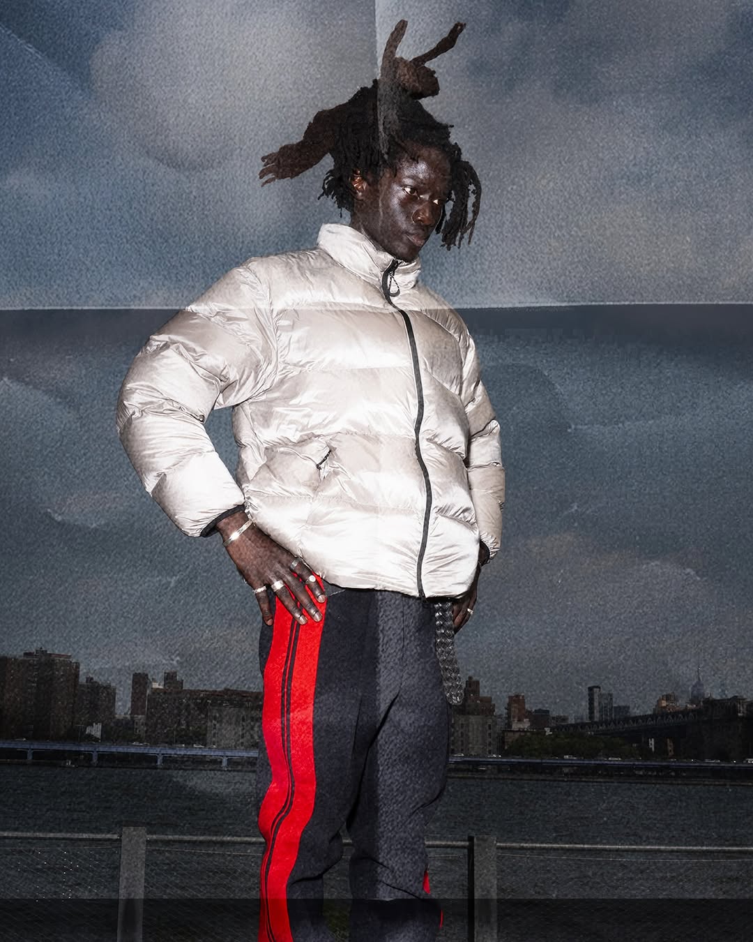 Portrait of an African American man wearing a white puffer jacket, dark pants, with a city skyline and cloudy sky in the backdrop.