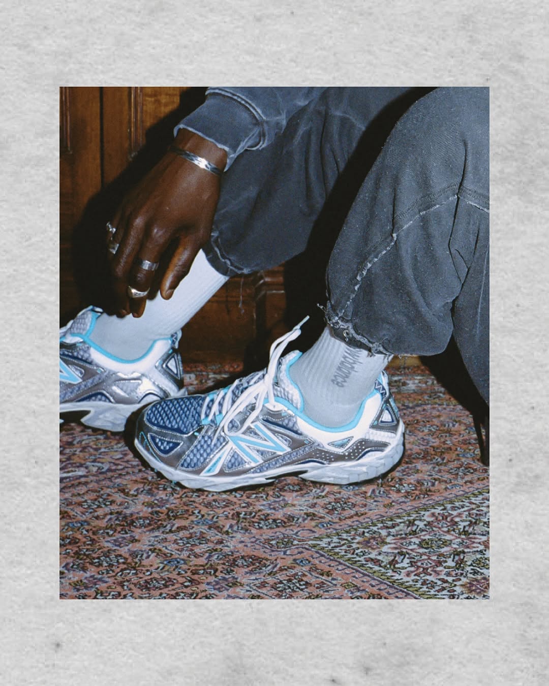 Eye-level studio shot of silver and blue New Balance running shoes on a detailed Persian-style rug; person seated on wooden surface.