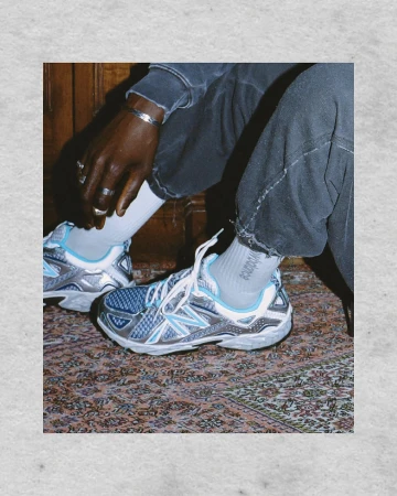 Eye-level studio shot of silver and blue New Balance running shoes on a detailed Persian-style rug; person seated on wooden surface.