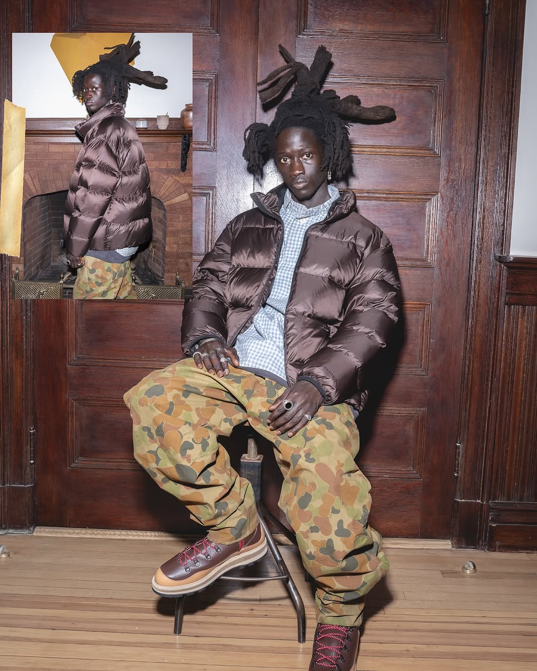 A young Black male model with dreadlocks sits indoors wearing a brown puffer jacket, patterned pants, and lug-sole shoes, in a styled portrait.