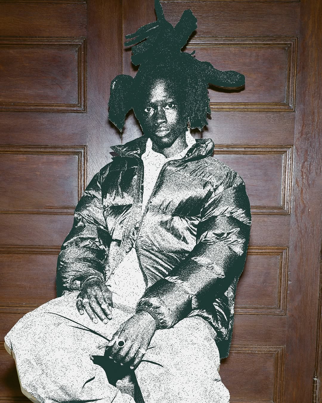 Portrait of a black adult male with an elaborate hairstyle wearing a puffer jacket indoors against a paneled wood wall.