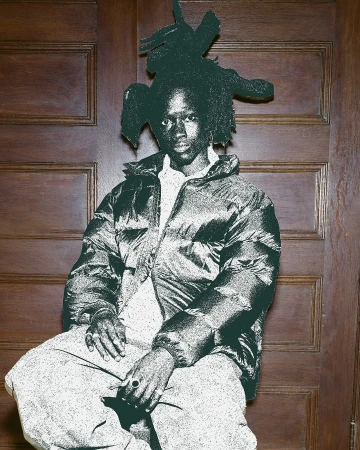 Portrait of a black adult male with an elaborate hairstyle wearing a puffer jacket indoors against a paneled wood wall.