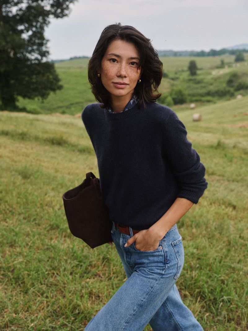 Portrait of an Asian woman in a blue sweater walking outdoors, carrying a tote bag in a grassy field with rolling hills.