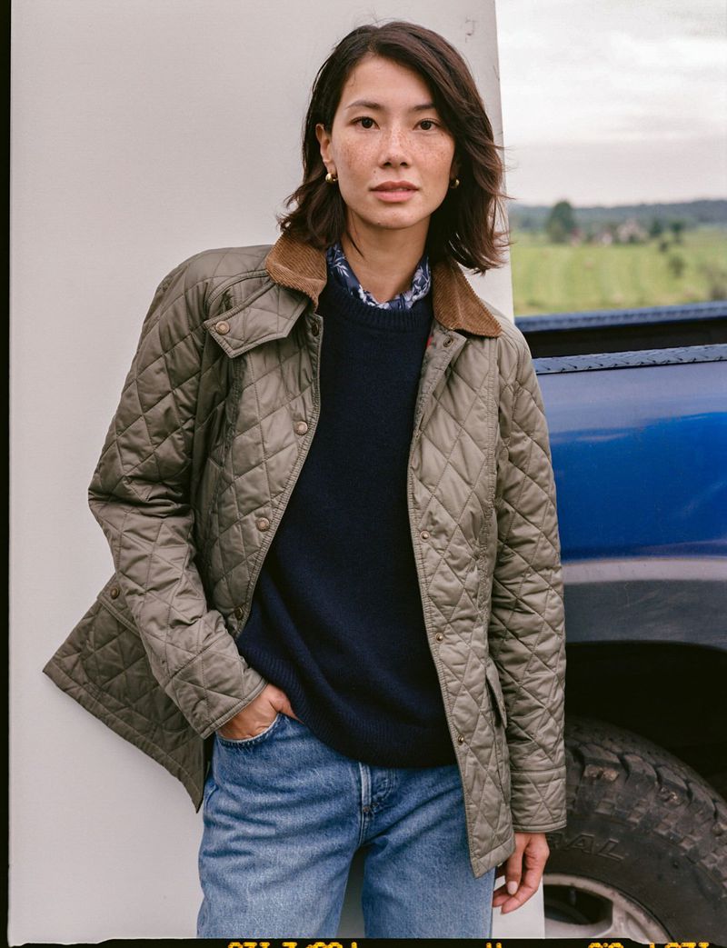 A woman in a quilted jacket, navy sweater, and jeans poses outdoors with a truck, showcasing a casual, stylish look in a natural setting.