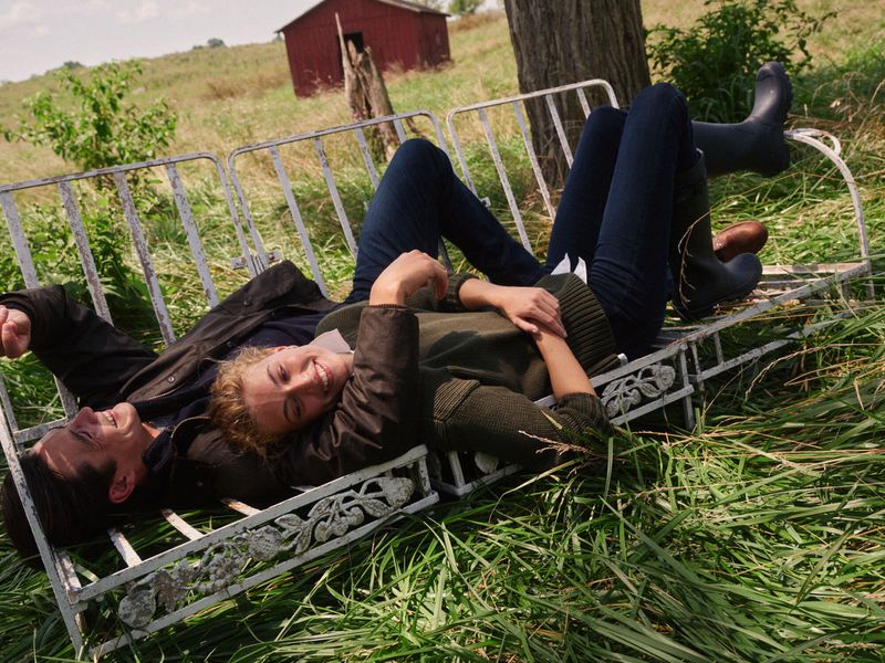 A happy couple reclines on an ornate metal bench amid a grassy field, sharing a lighthearted moment under natural light with a relaxed pose.