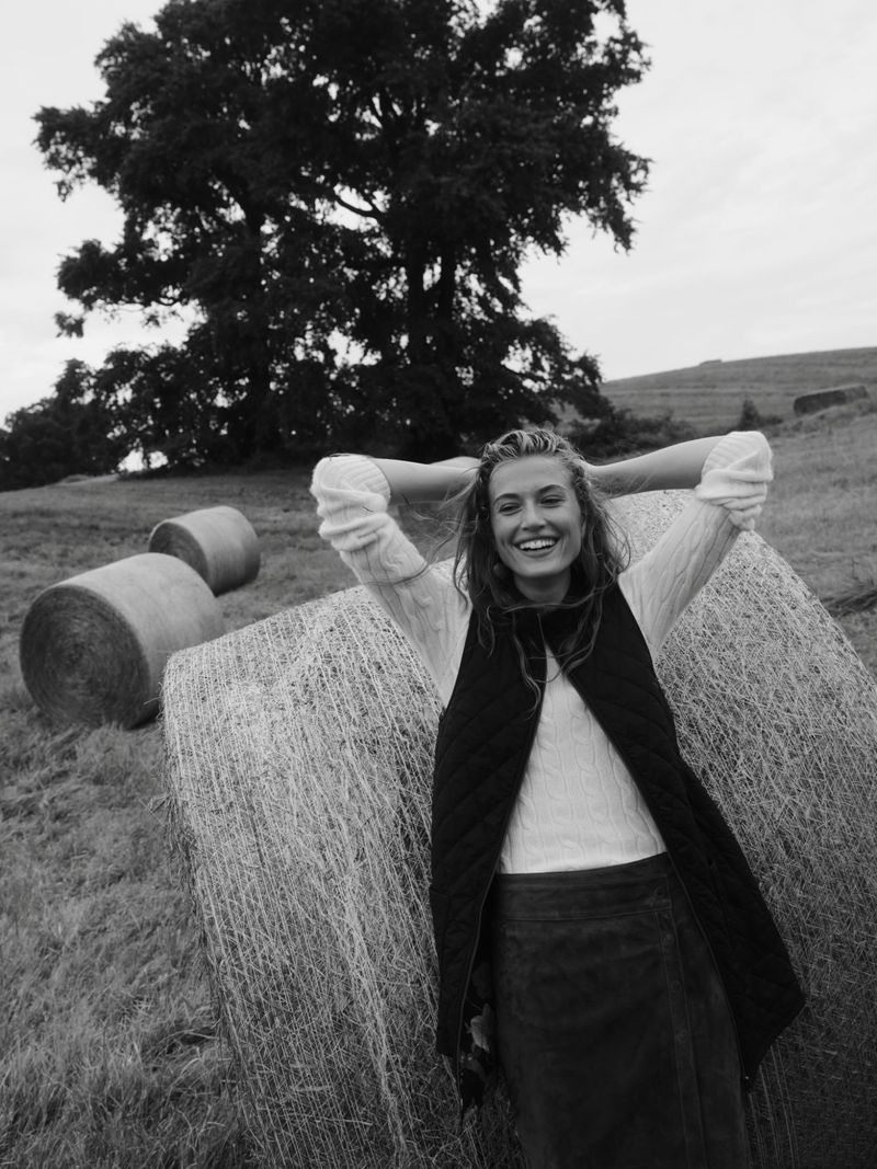 Black-and-white shot of smiling woman in vest and skirt standing by hay bales in a field, with hands behind her head.