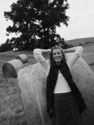 Smiling woman leaning against a large hay bale in a field wearing a quilted vest