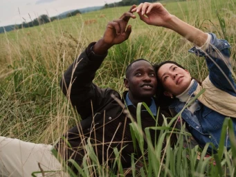 Two models lying in a tall grassy meadow reaching upward with hands gesturing towards the sky in an outdoor campaign shoot.