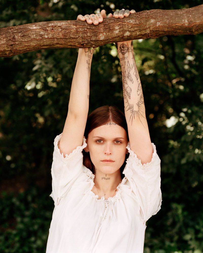 Portrait of a woman with arm tattoos, hanging from a tree branch in a white dress, standing in a green wooded outdoor area.