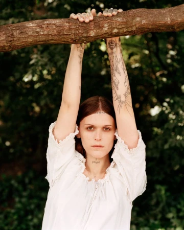 Portrait of a woman with arm tattoos, hanging from a tree branch in a white dress, standing in a green wooded outdoor area.