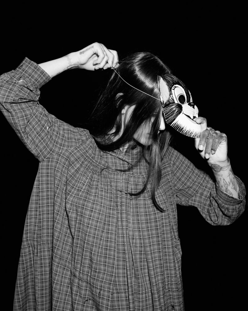 Person wearing a vintage-style plaid dress and placing a cartoon cat mask over their face in a monochrome shot against a dark background.