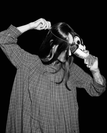 Person wearing a vintage-style plaid dress and placing a cartoon cat mask over their face in a monochrome shot against a dark background.